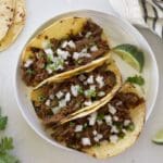 White plate with 3 Mexican shredded beef tacos and lime wedges.