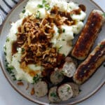 Plate of sliced Bangers and Mash topped with gravy and fried onions.